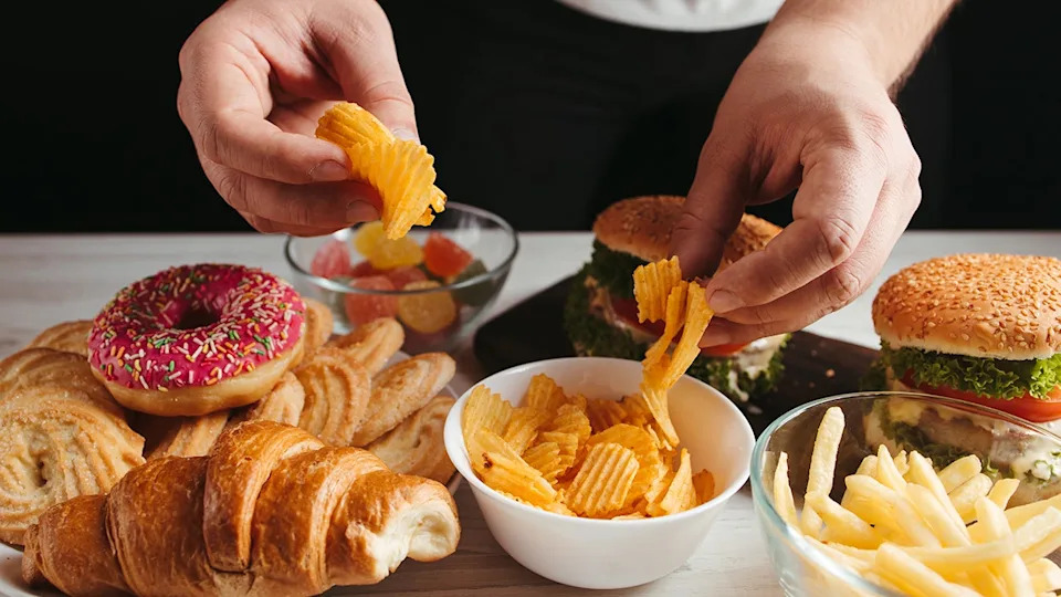 Man's hands reaching for unhealthy, processed snacks including chips, fries, packaged desserts, burgers and gummy candies.