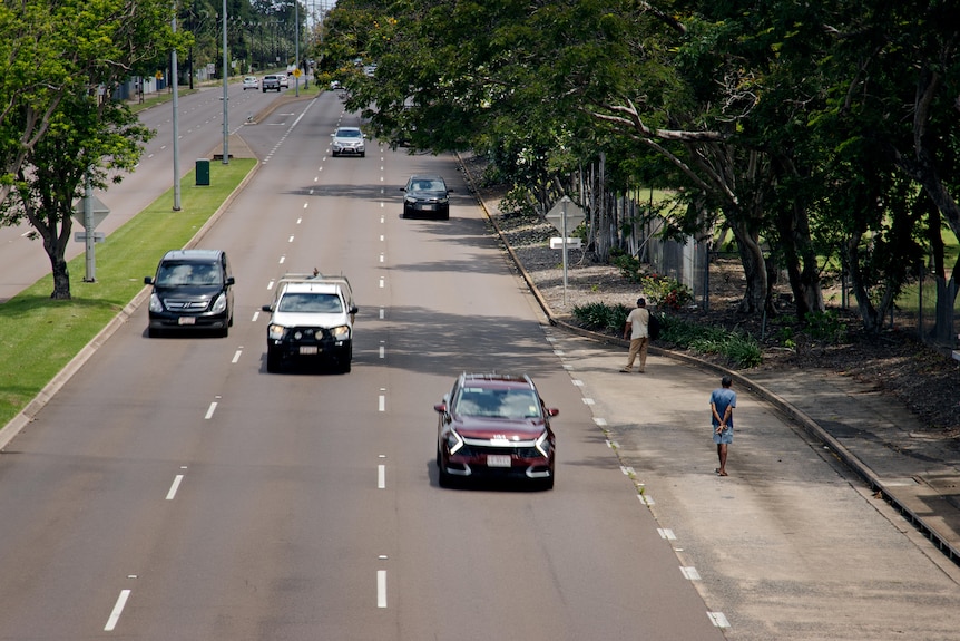 Cars on a main road. People are standing on the side of the road. 