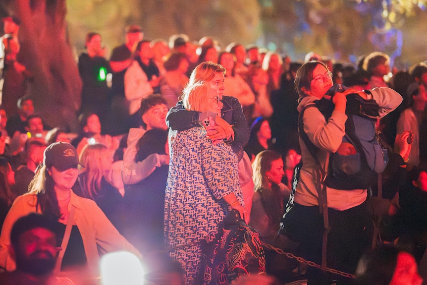 An older woman and a younger woman hug one another among a crowd of people watching the fireworks.