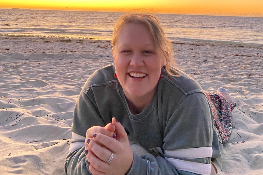 Woman with blonde hair wearing green jumper lies on beach with sunset in the background