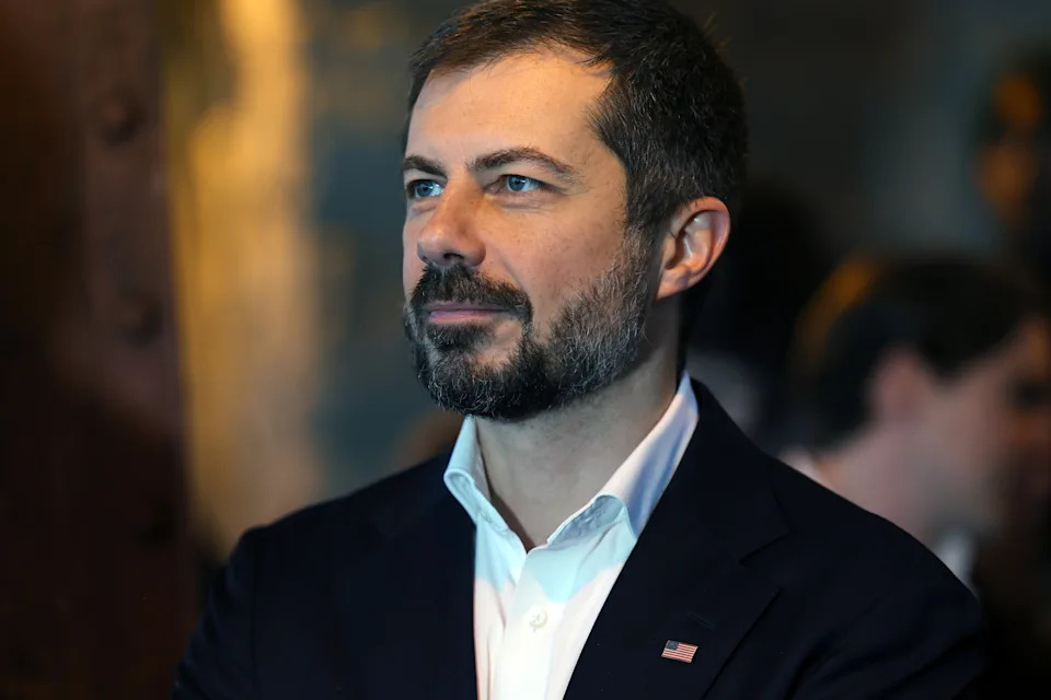 A person with a beard in a suit jacket adorned with a small flag pin gazes thoughtfully to the side