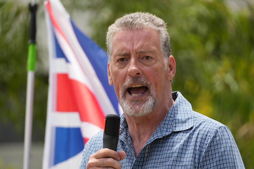 Stephen Wells speaks into a microphone, an Australian flag in the background.
