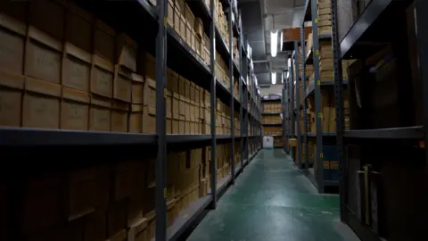 The photo looks down a green floored path in between steel racks from floor to ceiling which are filled with hundreds of brown cardboard boxes.