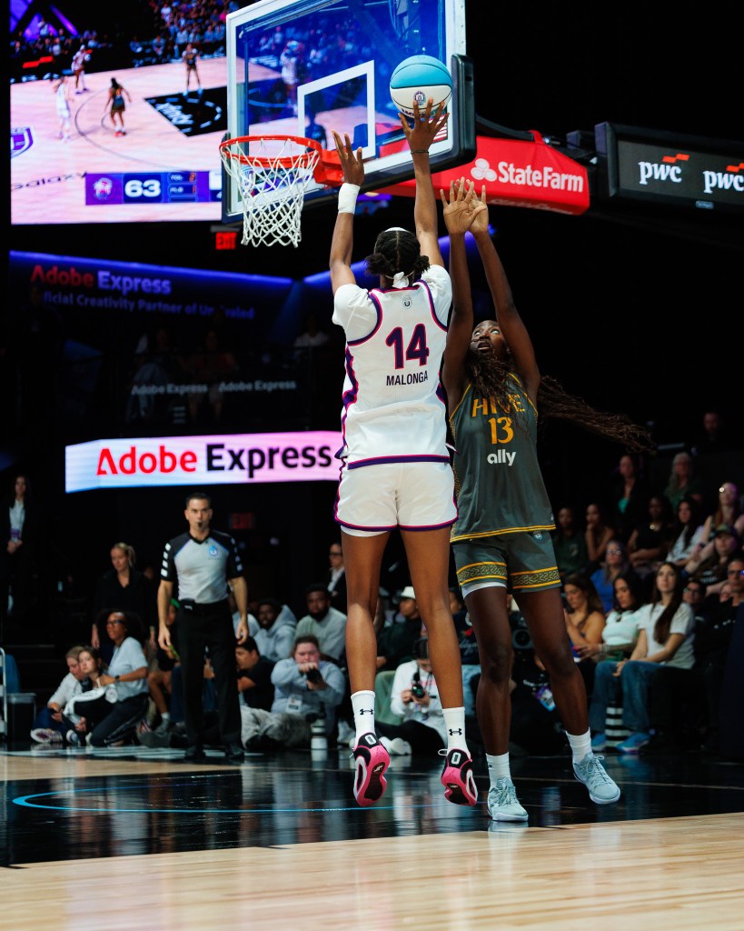 Two players jump for a ball on the court. One wears a white uniform and the other wears a green/gray uniform. 