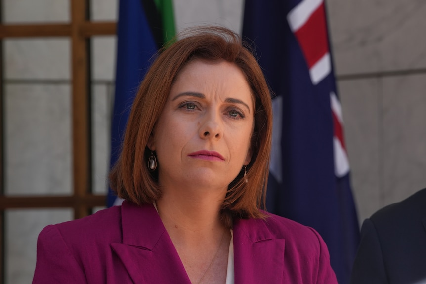 A red-haired woman in a blazer stands in front of an Australian flag.