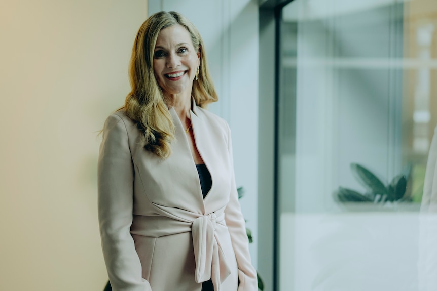 Julie smiles while standing next to a glass window wearing a beige suit jacket