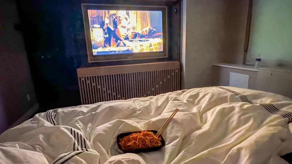 Takeout noodles with chopsticks on white bed with TV