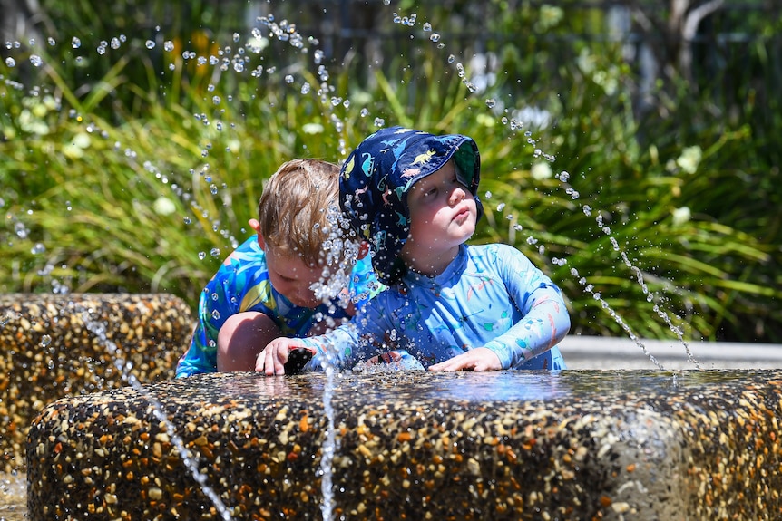 Toddlers wearing full-cover bathing suits and hats are playing in a fountain in Brisbane.