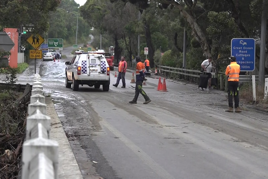 A police car and people in orange shirts and blue pants stand on a roadway covered in mud.