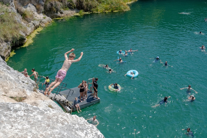 A boy in pink board shorts jumps from a rocky cliff towards a lake full of water, with swimmers below
