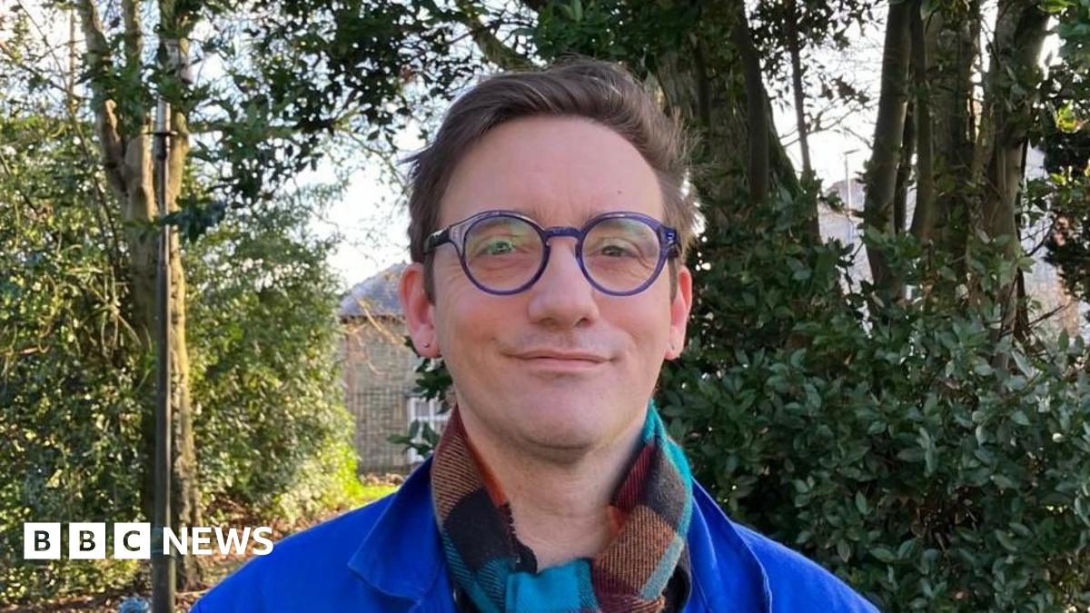 Shows a man in a blue jacket and checked scarf with round blue glasses standing in front of trees