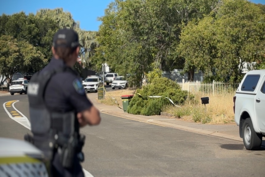 A police office arms crossed leaning on a police car looking down a street where police tape is across bushes in front of a hous