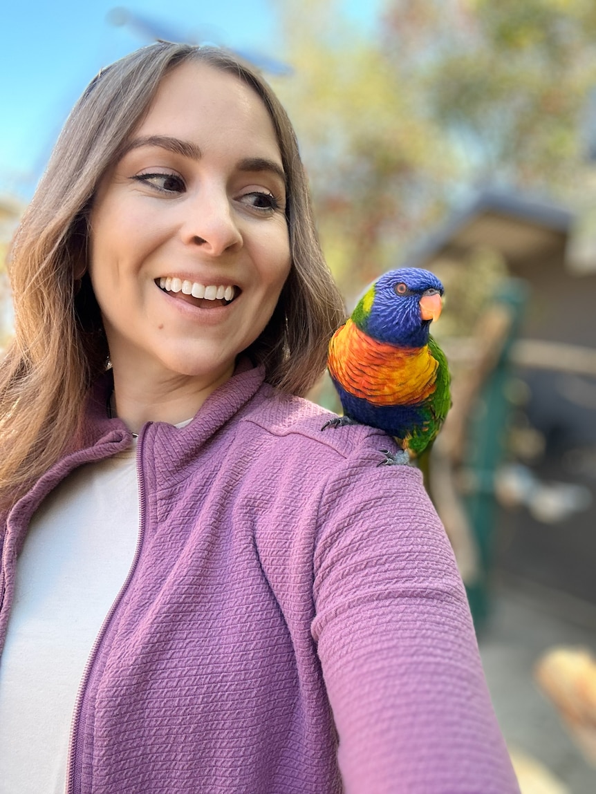 Woman wearing casuasl clothes satands with colourful bird on her shoulder