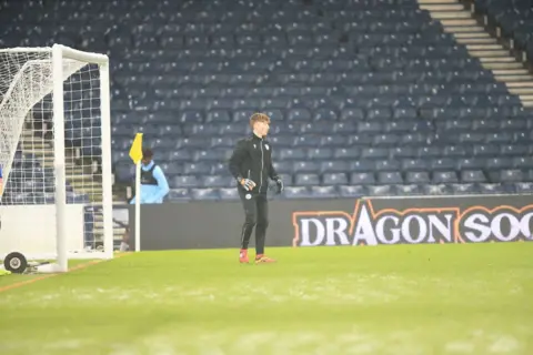 Queen of the South A goalkeeper in black kit stands in front of a set of goals with a large, empty stand behind him