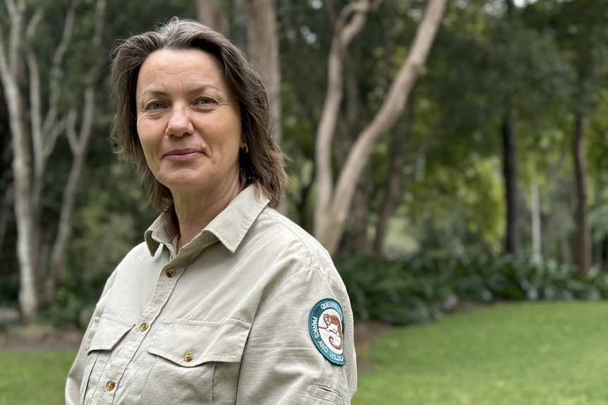 A woman wearing a khaki ranger shirt stands in a clearing near some bushland.