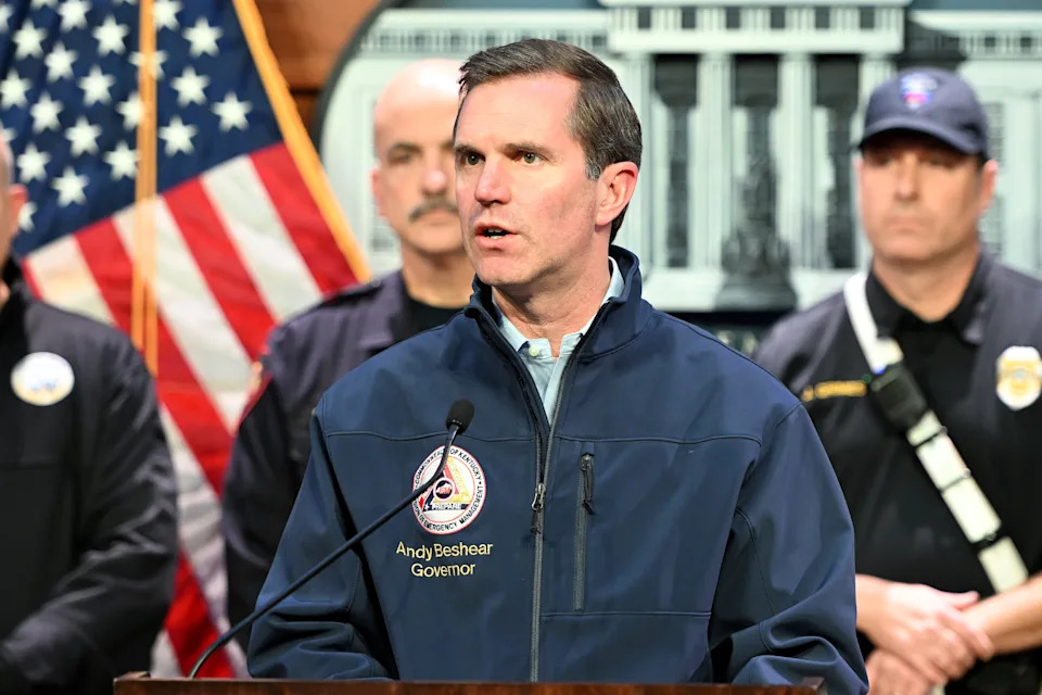 A man speaking at a podium with "Governor" on his jacket, flanked by uniformed personnel and an American flag in the background