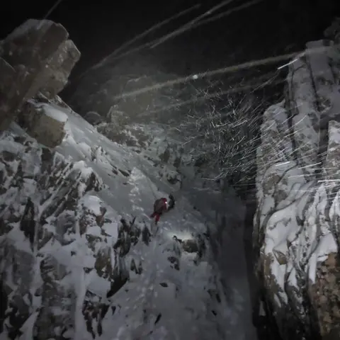 Glencoe Mountain Rescue Team A person climbing on a snowy rockface in the dark during a blizzard.