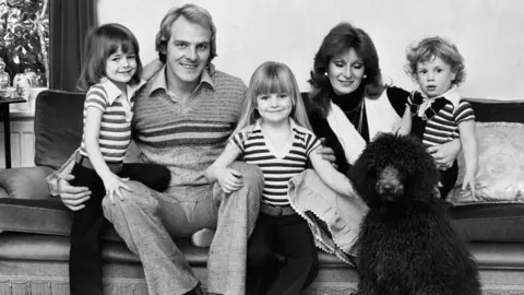 Getty Images A black and white image of a family of three children and a father and mother. All sat on a couch with a black fluffy dog.