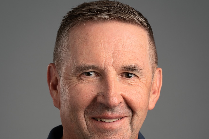 A middle aged man in a simple black polo looks at the camera, the background is grey. 