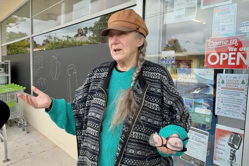 A photo of a woman with long gray hair in a plat, wearing a brown hat, blue jumper, standing on street