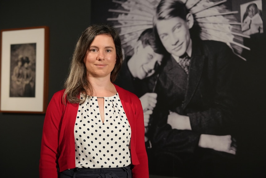 A woman looks at the camera standing in front of an old black and white photograph.