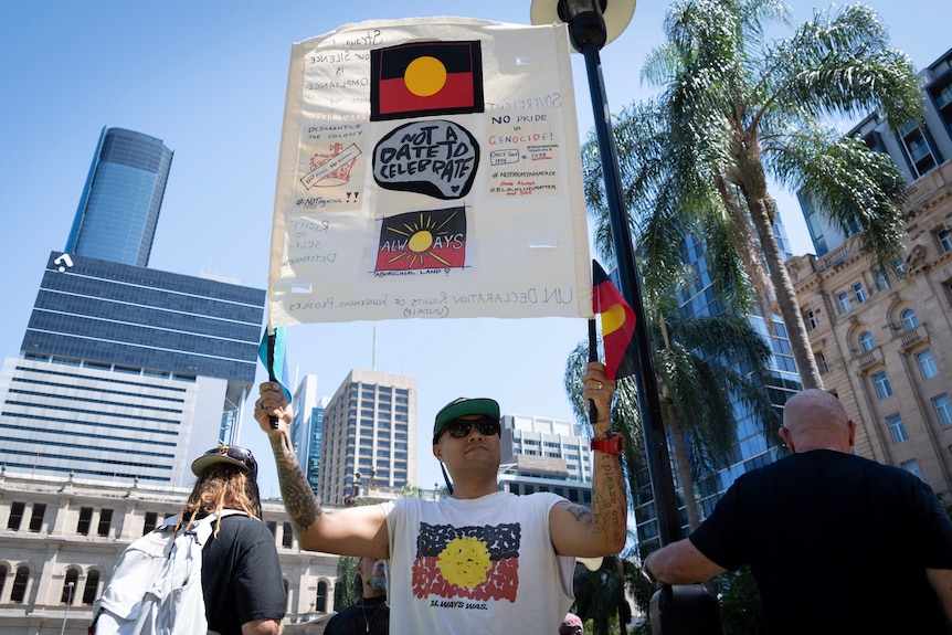 A man wearing a shirt featuring the Aboriginal flag holds up a sign.