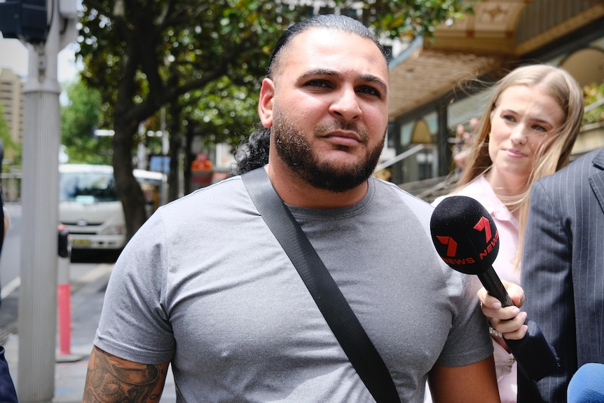 A close-up shot of a man with facial hair walking along a path surrounded by media and their microphones.