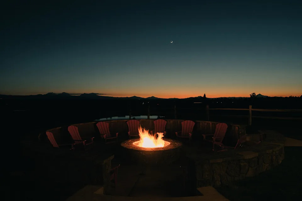 A fire in the center of the frame surrounded by dimly-lit chairs arranged in a circle around it. A nearly-dark landscape stretches out toward a sliver of orange sunset