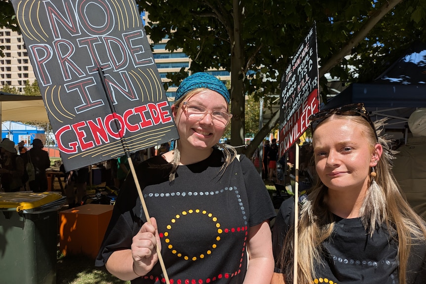 Two women dressed in shirts with the Aboriginal falg on them hold signs. One sign says "No pride in genocide".