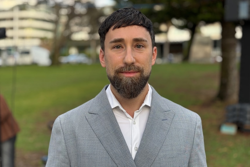 A dark-haired, bearded man in a sports coat stands in a park.
