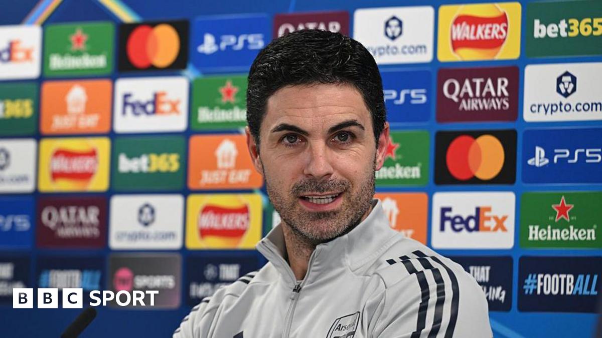 Mikel Arteta speaking at his news conference in a grey Arsenal tracksuit top, with a board of sponsor logos behind him