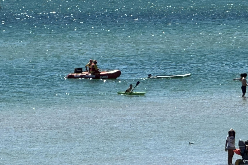 Two peple in an inflatable boat float behind a person whose head can be seen beside a paddle board on blue water.