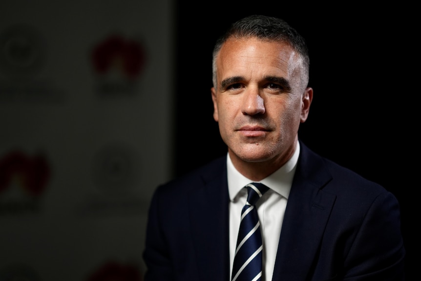 A close-up of a man with a serious expression looking directly into the camera. He wears a navy suit and tie and white shirt.