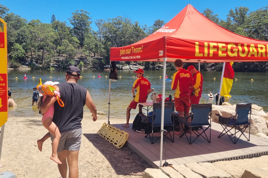 A man holding a girl walks towards the lake past a lifeguard tent with lifeguards keeping an eye on swimmers.