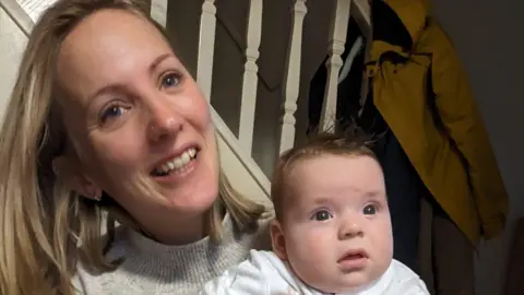 Louise smiles at the camera, holding her baby