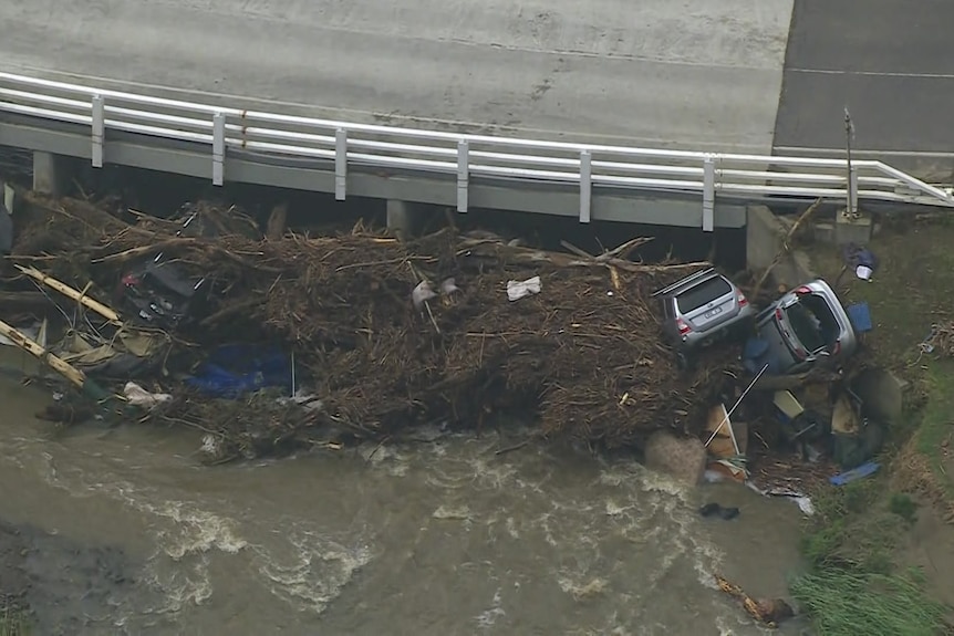 Two cars are half buried in a big pile of sticks and debris in water pushed up against a bridge.