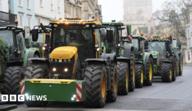 A queue of green and yellow tractors makes its way through an Oxford city centre road, with historic buildings in the background, under a misty sky