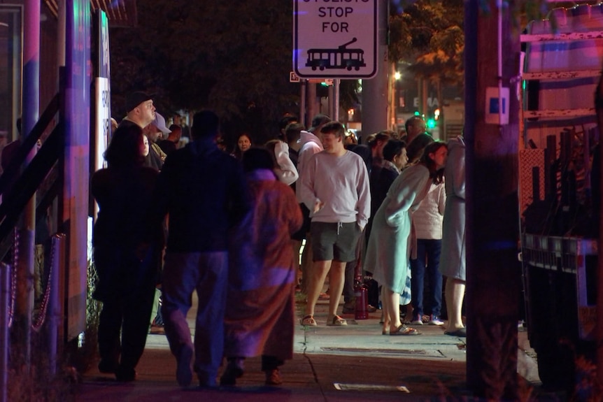 A large group of people stand on a footpath at night.