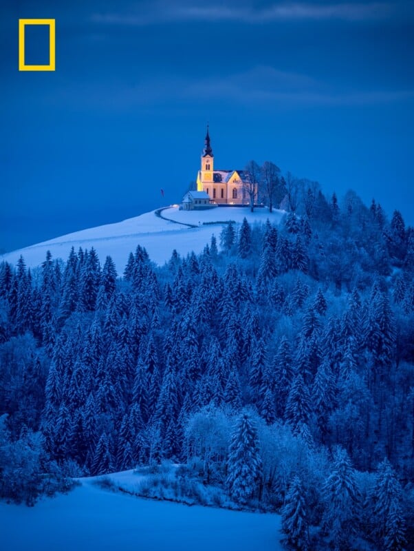 A warmly lit church sits atop a snowy hill, surrounded by dense, snow-covered forest under a blue twilight sky. A yellow National Geographic rectangle logo appears in the top left corner.