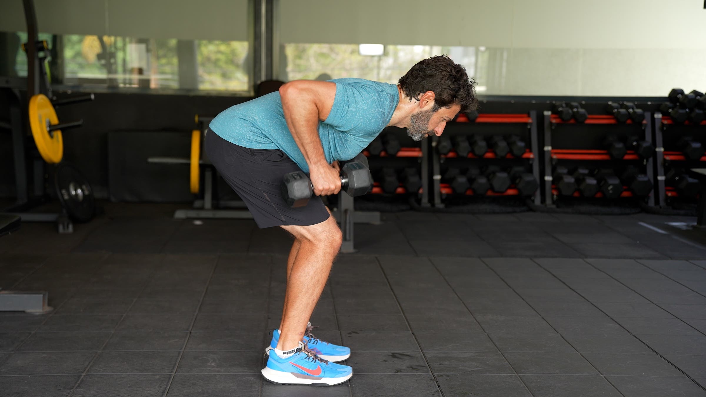 a man with dark hair wearing a blue shirt and black shorts does bent-over row with one dumbbell in each hand