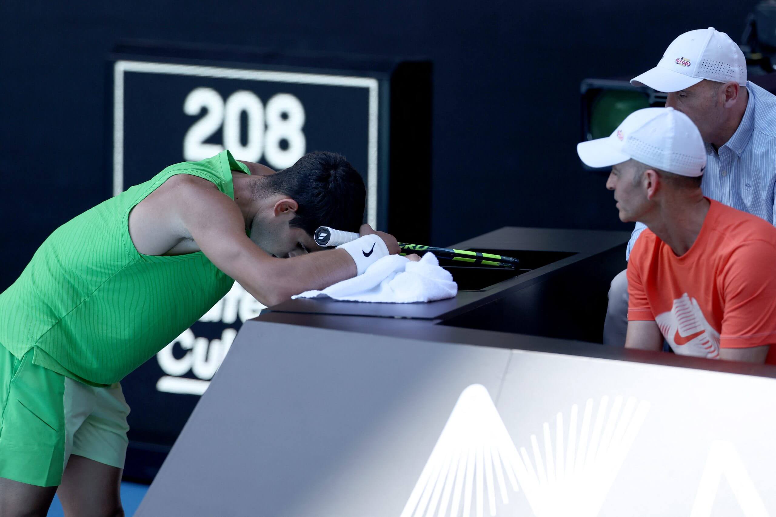 Carlos Alcaraz leans at the side of the court with his head in a towel in front of two of his coaches.