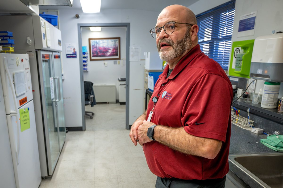A man in a red shirt stands in a laboratory or medical office, looking toward the camera, with lab equipment and supplies visible on counters and shelves.