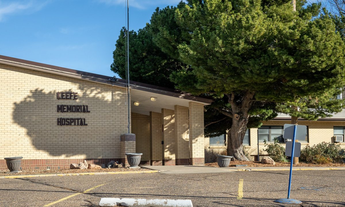 Exterior view of a tan brick building with the sign "Cee Vee Memorial Hospital" on the wall, large tree nearby, empty parking area in front.
