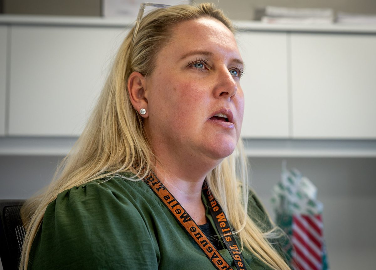 A woman with long blonde hair, wearing a green top and an orange lanyard, sits in an office environment with shelves and documents in the background.