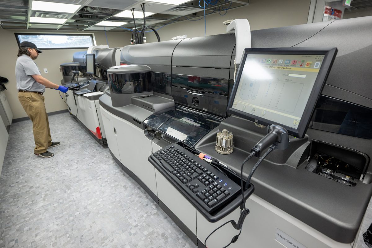A laboratory technician operates large automated analyzers in a modern lab, with a computer and barcode scanner visible in the foreground.