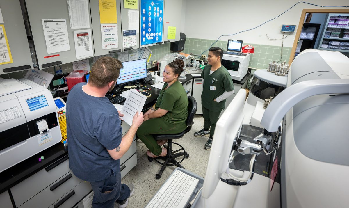 Three healthcare professionals are working together in a hospital laboratory, surrounded by computers, medical equipment, and documents.