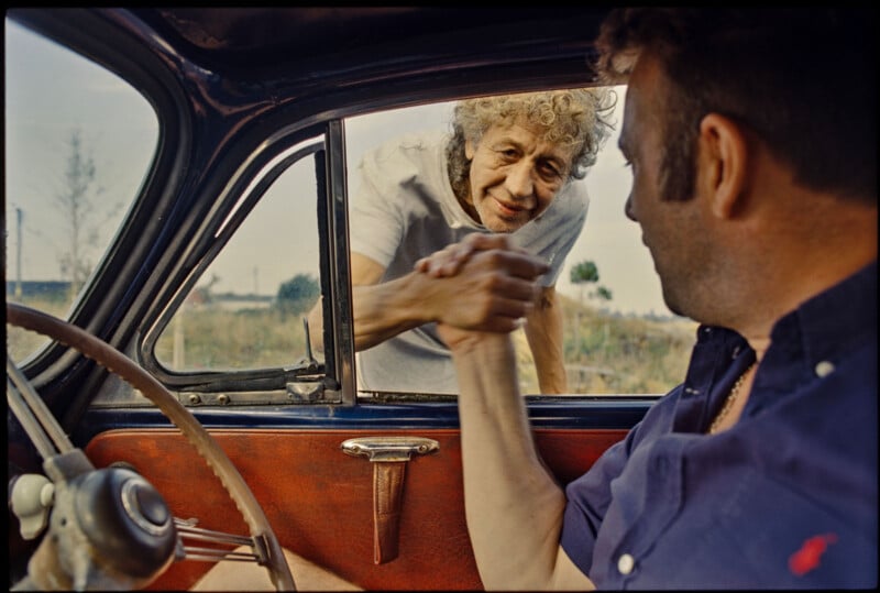 An older woman leans into an open car window, smiling and clasping hands with a man seated inside the car, creating a warm and friendly connection between them.