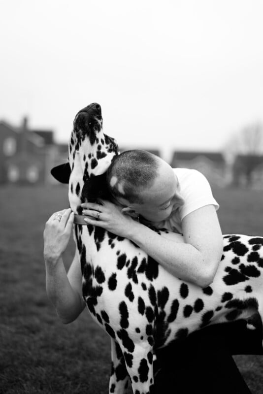A person with a shaved head hugs a Dalmatian dog outdoors. The person smiles, pressing their face against the dog's neck. The background is blurred with houses and grass visible. The image is in black and white.