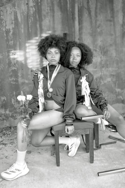 Two young women sit side by side on a wooden bench, wearing athletic jackets adorned with medals and ribbons. One holds white roses. Both have natural curly hair and serious expressions, against a textured wall background.
