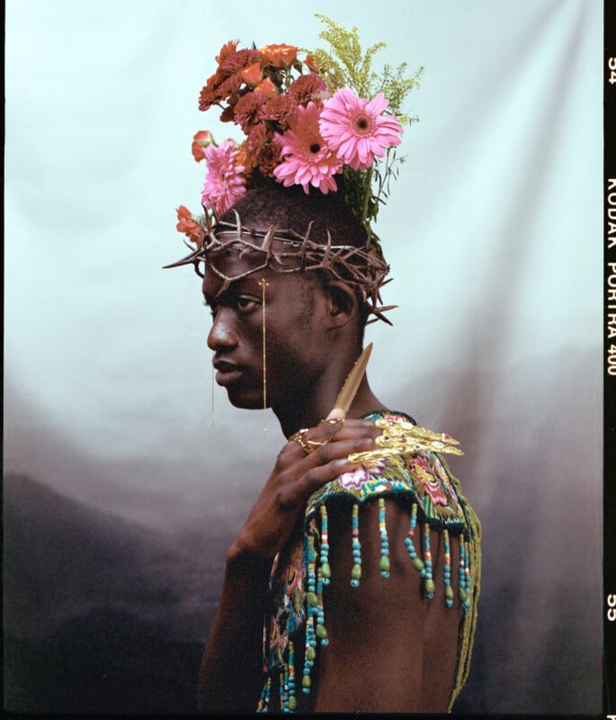A person in profile wears a crown of thorns and a floral headpiece with pink and red flowers. They are dressed in an ornate, beaded garment against a soft, gradient background.